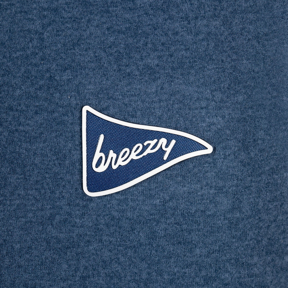 A close-up of a heathered blue fabric surface with a small embroidered pennant-shaped logo in blue and white reading 'breezy'.