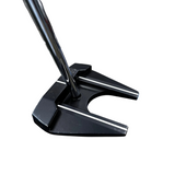 A close-up of a black golf putter head with a silver metal shaft, photographed in a golf shop setting with a blurred background and a blue bag on the right.