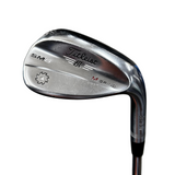 A close-up shot of a Titleist Bv SM6 M Grind golf iron club head, showing the polished metal finish and engraved branding, with the club shaft extending downward against a blurred gray concrete background.