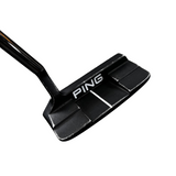 A close-up shot of a black Ping golf putter head with silver lettering, resting on a gray concrete floor with store displays blurred in the background.