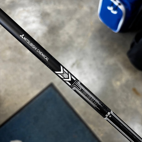 A close-up diagonal shot of a black Mizubishi Chemical golf club shaft with white branding, set against a concrete floor background with a blue bag and mat partially visible.