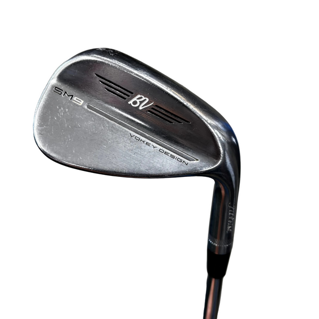 A close-up shot of a titleist SM9 BV golf iron wedge, showing the club head and part of the shaft against a gray concrete floor background.
